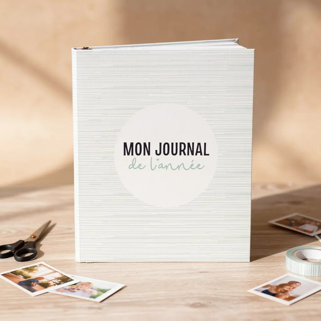 Album photo blanc 23x30 cm intitulé “Mon journal de l'année - 2026” posé sur une table avec pellicule et photos imprimées autour.